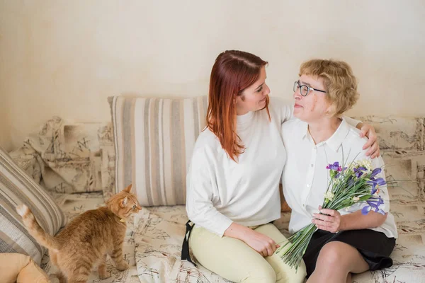 Η κόρη δίνει λουλούδια σε μια ενήλικη μητέρα που κάθεται στον καναπέ στο σαλόνι. Περνάμε χρόνο μαζί, γιορτάζοντας στο σπίτι τα Σαββατοκύριακα. Ημέρα της μητέρας. Θερμές σχέσεις μεταξύ των γενεών — Φωτογραφία Αρχείου