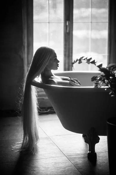 Eine schöne Frau mit wunderschönen langen blonden Haaren entspannt in der Badewanne. Silhouette einer Frau im Profil, die im Badezimmer liegt. — Stockfoto