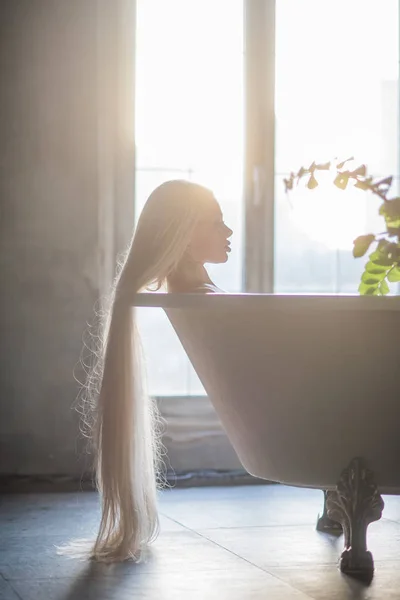 Una bella donna con splendidi lunghi capelli biondi è rilassante nella vasca da bagno. Silhouette di una donna di profilo sdraiata in bagno . — Foto Stock