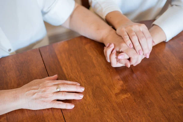 Elderly mother and her daughter holding hands while sitting at the table.Close up on women of different generations holding hands. Close Up Shot Of Mother And Daughter\'s Hands Holding