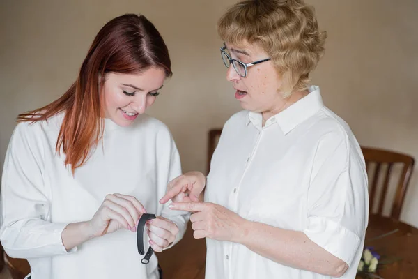 A woman helps an older mother to put on a fitness bracelet. An adult daughter helps her mother to sort out the device. Woman helps grandmother to set up a smart watch