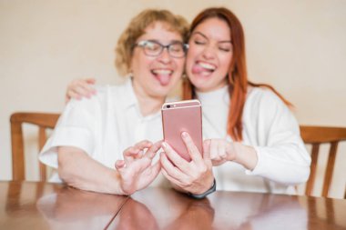 Anne ve kızı masada evde otururken akıllı telefonun ön kamera fotoğraflandı. Neşeli yaşlı anne ve kızı komik selfie'ler yapıyor ve dillerini gösteriyor.