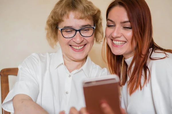 Una anciana madre e hija vestidas con ropa ligera sonríen y se toman selfies. Mamá e hija son fotografiadas en la cámara frontal del teléfono inteligente mientras están sentadas en casa en la mesa . — Foto de Stock