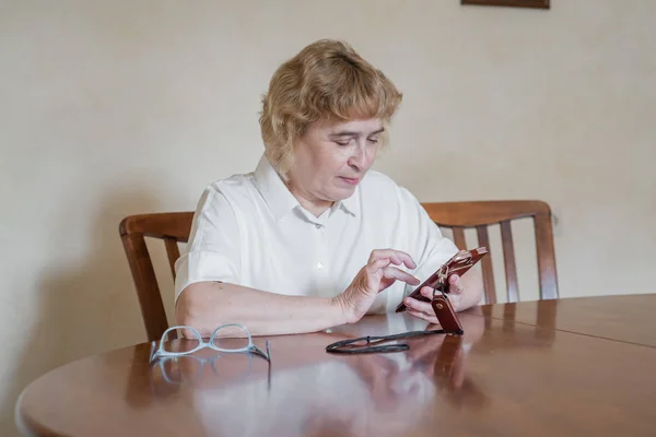 Hermosa anciana en una blusa blanca sentada a la mesa y escribe mensajes en el teléfono. Mujer jubilada anotó puntos y lee el mensaje en el teléfono inteligente — Foto de Stock