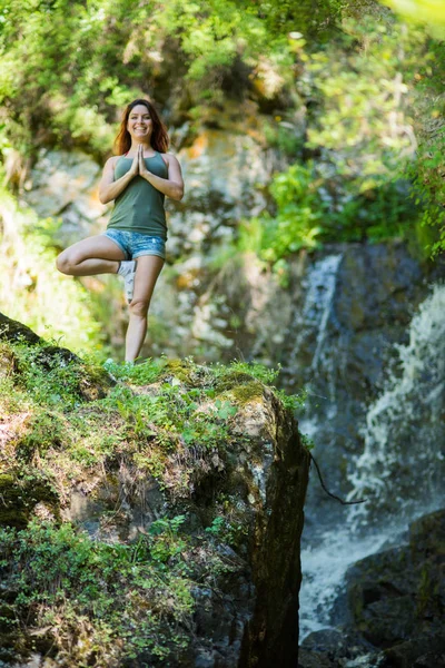Red-haired woman in shorts doing yoga at the waterfall. A girl stands in a pose of a tree in the forest — Stock Photo, Image