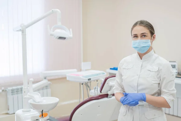 Attraente medico donna in uniforme bianca sul posto di lavoro. Una giovane bella dentista in maschera e guanti è in piedi nello studio vicino alla sedia — Foto Stock