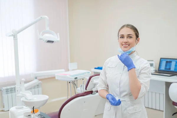 Attraente sorridente medico donna in uniforme bianca sul posto di lavoro. Una giovane bella dentista in maschera e guanti è in piedi nello studio vicino alla sedia — Foto Stock