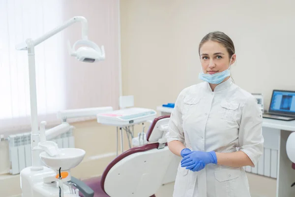 Приваблива усміхнена жінка-лікар у білій формі на робочому місці. Молода красива жінка-стоматолог в масці і рукавичках стоїть в офісі біля стільця — стокове фото