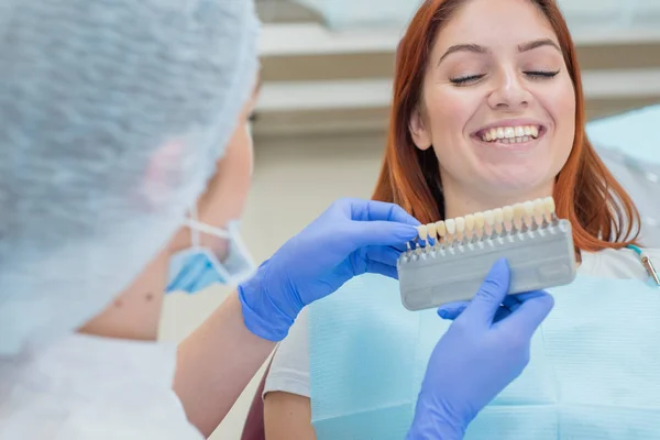 Verifique e selecione a cor dos dentes na cadeira do dentista. Dentista faz o processo de tratar uma bela jovem ruiva. Seleção de um implante dentário . — Fotografia de Stock