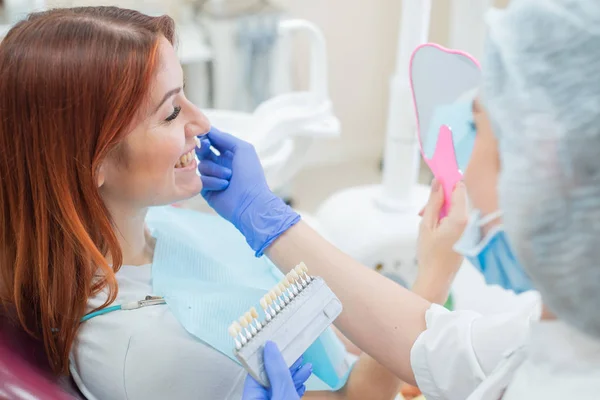 Compruebe y seleccione el color de los dientes en la silla del dentista. Dentista hace el proceso de tratar a una hermosa chica pelirroja joven. Selección de un implante dental. Paciente mirándose en el espejo . — Foto de Stock