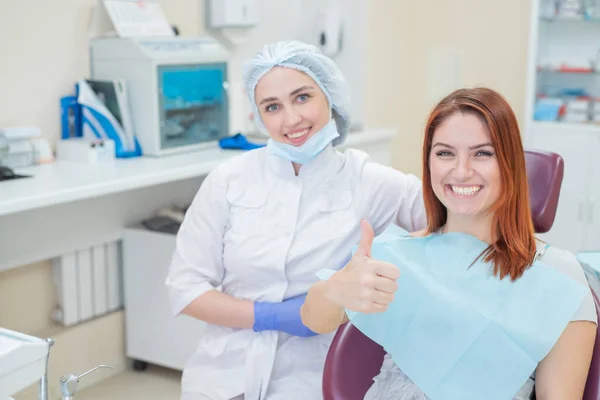 快乐的妇女牙医和病人在牙科办公室治疗牙齿后，面带微笑，看着相机。快乐的病人把拇指当作背书。牙科设备. — 图库照片