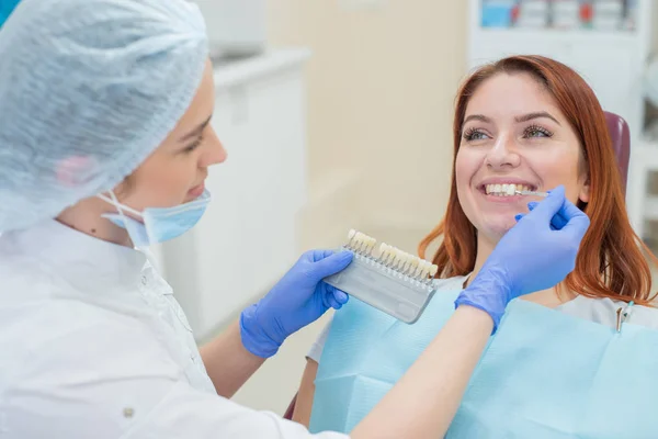 Controllare e selezionare il colore dei denti sulla sedia del dentista. Il dentista fa il processo di trattamento di una bella giovane ragazza dai capelli rossi. Selezione di un impianto dentale . — Foto Stock