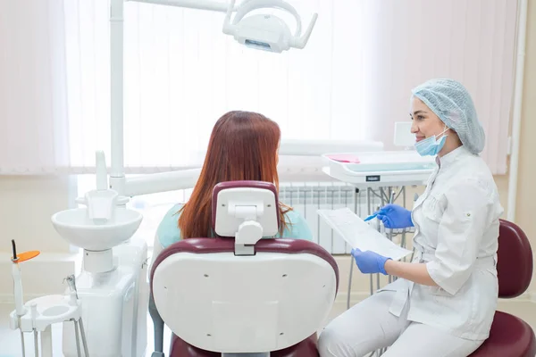 年轻漂亮的女牙医在办公室与病人交谈。一位红头发的妇女坐在椅子上,坐着正畸医生,听预约。牙科服务。从背面查看 — 图库照片