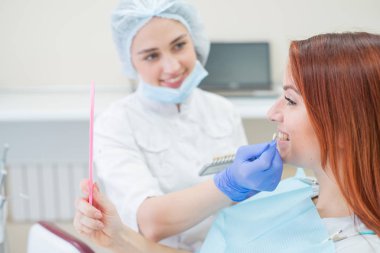 Diş hekimleri sandalyesinde dişlerin rengini kontrol edin ve seçin. Diş hekimi güzel bir genç kızıl saçlı kız tedavi süreci yapar. Bir diş implantı seçimi. Hasta aynaya bakıyor.