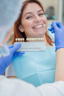Diş hekimi sandalyesinde dişlerin rengini kontrol edin ve seçin. Diş hekimi güzel bir genç kızıl saçlı kız tedavi süreci yapar. Diş implantının seçimi.