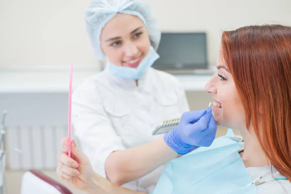 Controllare e selezionare il colore dei denti nella sedia dentista. Il dentista fa il processo di trattamento di una bella ragazza dai capelli rossi. Selezione di un impianto dentale. Il paziente si guarda allo specchio . — Foto Stock