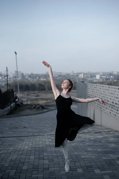 Ballerina in a tutu posing next to the fence. Beautiful young woman in black dress and pointe dancing over city background. Portrait of a gorgeous ballerina performing a dance outdoors — Stock Photo, Image