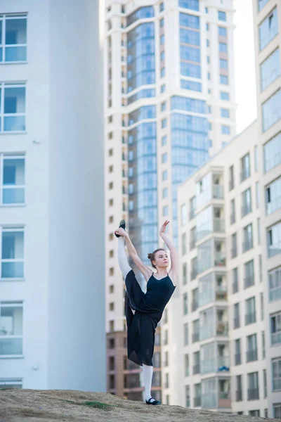 Ballerina in tutù posa contro un edificio residenziale. Bella giovane donna in un abito nero e scarpe da punta salto dimostra stretching. La ballerina è in piedi tra le spaccature. Fuori. . — Foto Stock