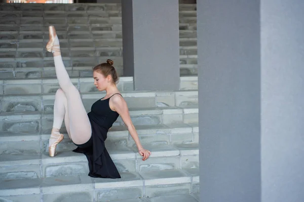 Балерина в Туту позують сидячи на сходах красивою молодою жінкою в чорному платті і Пуант черевики сидять в елегантній позі на сходах зовні — стокове фото