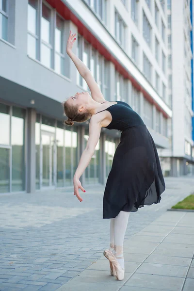 Bailarina em um tutu posando na frente de um edifício residencial de vários andares. Mulher bonita em vestido preto e sapatos pontiagudos com incrível flexibilidade. Linda bailarina executa um ba elegante — Fotografia de Stock