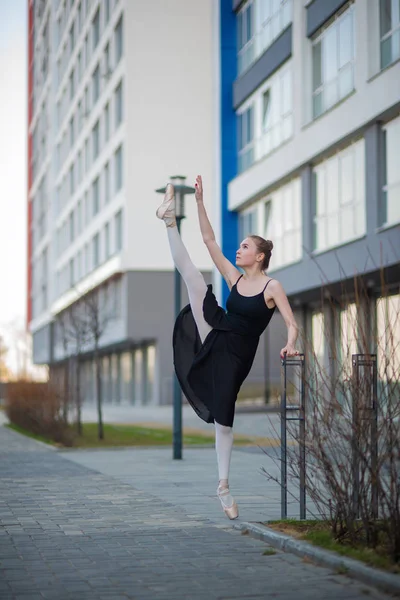 Ballerina in a tutu posing against the backdrop of a residential building. A beautiful young woman in a black dress and pointe shoes demonstrates an excellent stretch. Ballerina is standing in the spl — Stock Photo, Image