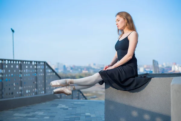 Ballerina in tutu posing sitting. Beautiful young woman in black dress and pointe dancing over city background. Portrait of a gorgeous ballerina performing a dance outdoors — Stock Photo, Image