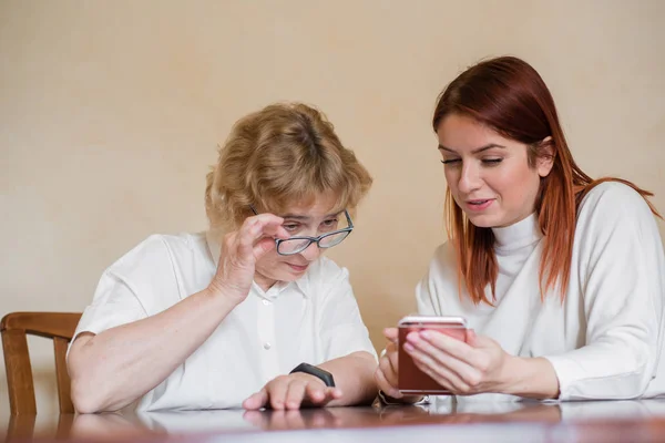 Imagen interior de una hermosa joven pelirroja sosteniendo un teléfono móvil, mostrando a su atractiva madre anciana cómo usar un gadget electrónico. Chica mostrar cómo utilizar el teléfono inteligente a su madre . — Foto de Stock