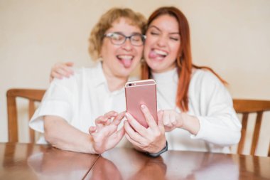 Anne ve kızı masada evde otururken akıllı telefonun ön kamera fotoğraflandı. Neşeli yaşlı anne ve kızı komik selfie'ler yapıyor ve dillerini gösteriyor.
