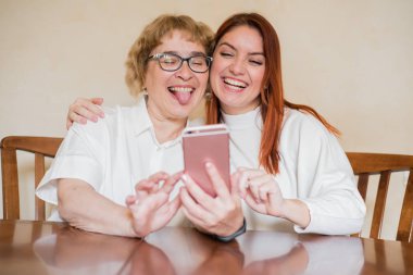 Anne ve kızı masada evde otururken akıllı telefonun ön kamera fotoğraflandı. Neşeli yaşlı anne ve kızı komik selfie'ler yapıyor ve dillerini gösteriyor.