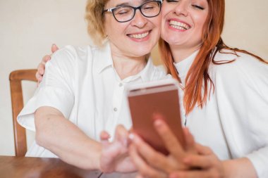 Hafif kıyafetler giymiş yaşlı bir anne ve kızı gülümsüyor ve selfie çekiyor. Anne ve kızı masada evde otururken akıllı telefonun ön kamera fotoğraflandı.