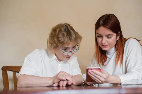 Imagen interior de una hermosa joven pelirroja sosteniendo un teléfono móvil, mostrando a su atractiva madre anciana cómo usar un gadget electrónico. Chica mostrar cómo utilizar el teléfono inteligente a su madre . — Foto de Stock