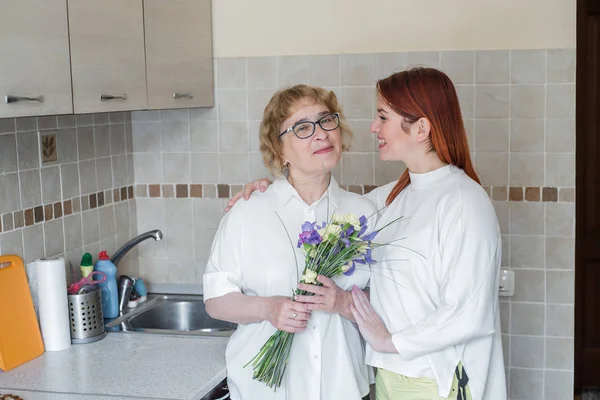 Lánya ad virágot egy felnőtt anya és ölelést neki a konyhában. Időtöltést együtt, ünneplő otthon a hétvégén. Anyák napja — Stock Fotó