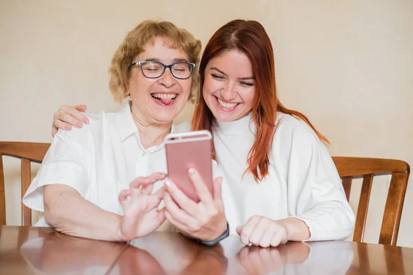 Anne ve kızı masada evde otururken akıllı telefonun ön kamera fotoğraflandı. Neşeli yaşlı anne ve kızı komik selfie'ler yapıyor ve dillerini gösteriyor. — Stok fotoğraf