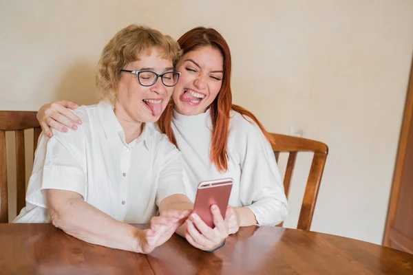 Anne ve kızı masada evde otururken akıllı telefonun ön kamera fotoğraflandı. Neşeli yaşlı anne ve kızı komik selfie'ler yapıyor ve dillerini gösteriyor. — Stok fotoğraf