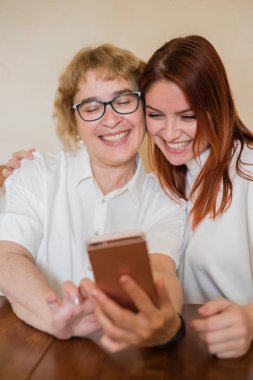 Hafif kıyafetler giymiş yaşlı bir anne ve kızı gülümsüyor ve selfie çekiyor. Anne ve kızı masada evde otururken akıllı telefonun ön kamera fotoğraflandı.