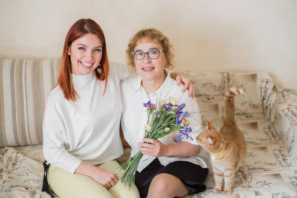 Η κόρη δίνει ένα μπουκέτο σε μια ενήλικη μητέρα που κάθεται σε έναν καναπέ στο σαλόνι. Περνάμε χρόνο μαζί, γιορτάζοντας στο σπίτι τα Σαββατοκύριακα. Ημέρα της μητέρας. Η γάτα τρώει λουλούδια — Φωτογραφία Αρχείου