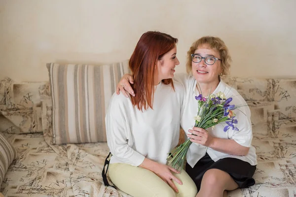 Η κόρη δίνει λουλούδια σε μια ενήλικη μητέρα που κάθεται στον καναπέ στο σαλόνι. Περνάμε χρόνο μαζί, γιορτάζοντας στο σπίτι τα Σαββατοκύριακα. Ημέρα της μητέρας. Θερμές σχέσεις μεταξύ των γενεών — Φωτογραφία Αρχείου