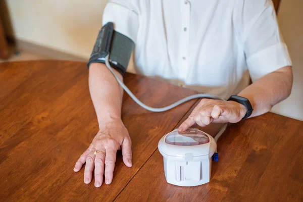 Μια ηλικιωμένη γυναίκα μετρά την αρτηριακή πίεση με μια ηλεκτρική συσκευή. Ένας συνταξιούχος χρησιμοποιεί ένα τόμετρο. Τα χέρια μιας παλιάς γυναίκας — Φωτογραφία Αρχείου