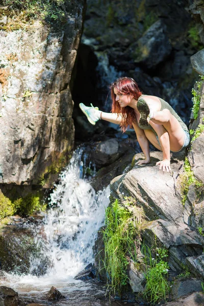 Red-haired woman in shorts doing yoga at the waterfall. The girl is standing in the pose of a firefly, titibhasana — Stock Photo, Image