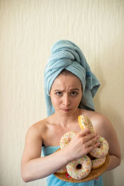 La chica en una toalla desayuna en la cama. Una joven atractiva come rosquillas en la cama. La chica con una toalla en la cabeza come rosquillas alegremente. . —  Fotos de Stock