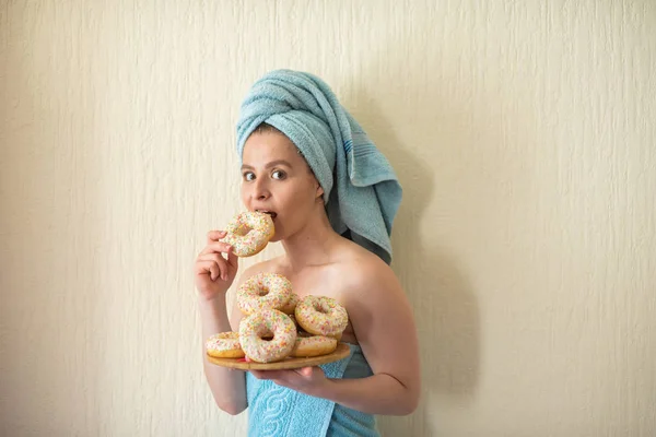 La fille dans une serviette prend le petit déjeuner dans le lit. Une jeune femme séduisante mange des beignets au lit. La fille avec une serviette sur la tête mange joyeusement des beignets . — Photo