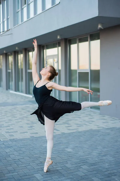 Bailarina en un tutú posando sobre el telón de fondo de un edificio residencial. Hermosa mujer joven en vestido negro y zapatos puntiagudos bailando ballet afuera . —  Fotos de Stock