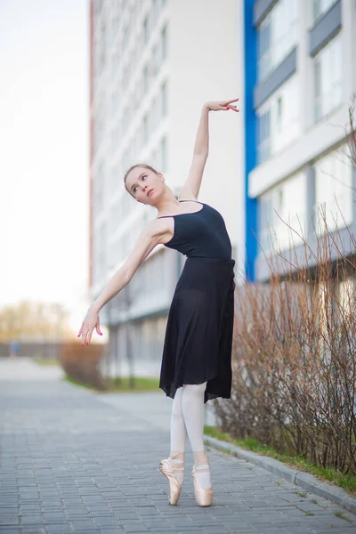 Bailarina en un tutú posando sobre el telón de fondo de un edificio residencial. Hermosa joven en vestido negro y zapatos puntiagudos bailando ballet al aire libre. Danza clásica elegante —  Fotos de Stock