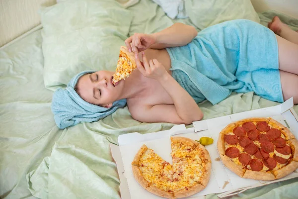 Ένα χαριτωμένο κορίτσι με μια πετσέτα στο κεφάλι της τρώει πίτσα στο κρεβάτι. Μια νεαρή γυναίκα που τρώει πίτσα στο κρεβάτι. Η ζωή είναι χαρά, σώμα θετικό. Αγάπη για το ιταλικό φαγητό. Διατροφικές συνήθειες, εθισμός σε φαστ φουντ. — Φωτογραφία Αρχείου