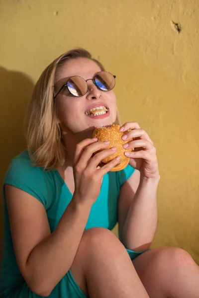 Šťastná žena v tyrkysově šatech sedí na chodníku a sní Burger v teplém letním dnu. Krásná blondýnka v slunečních brýlích má na žluté stěně obytné budovy podřadné pokrmy. — Stock fotografie