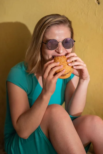 Щаслива жінка в бірюзовій сукні сидить на тротуарі і їсть бургер в теплий літній день. Красива блондинка в сонцезахисних окулярах насолоджується нездоровою їжею на жовтій стіні житлового будинку . — стокове фото