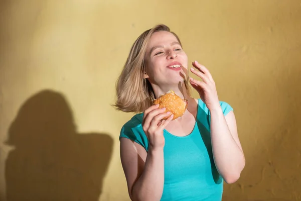 Портрет милої дівчини в зеленій сукні, що виходить на вулицю і їсть бургер в теплий літній день. Красива блондинка насолоджується фаст-фудом у жовтій стіні надворі. Неправильні звички їжі . — стокове фото