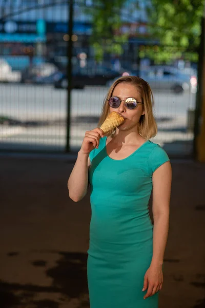 Счастливая женщина в бирюзовом платье стоит во дворе и ест вафельный рожок в теплый летний день. Красивая блондинка в солнечных очках наслаждается мороженым во время ходьбы. Летний десерт. — стоковое фото