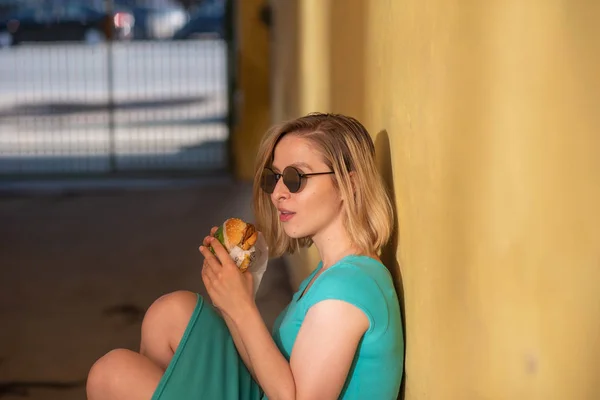 Uma mulher feliz em um vestido turquesa senta-se na calçada e come um hambúrguer em um dia quente de verão. Loira bonita em óculos de sol goza de junk food na parede amarela de um edifício residencial . — Fotografia de Stock