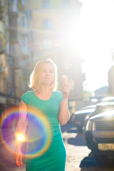 Retrato de una linda chica en un vestido verde camina por el patio y come helado. Una hermosa mujer rubia está comiendo postre afuera en un hermoso día de verano. La vida es un placer vacaciones —  Fotos de Stock